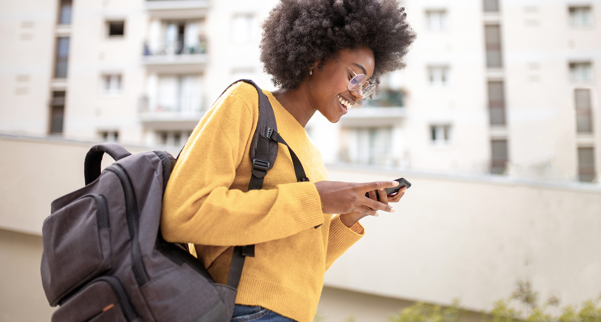 persoon met gele trui, bril met draadrand en grijze rugzak die buiten naar een telefoonscherm lacht