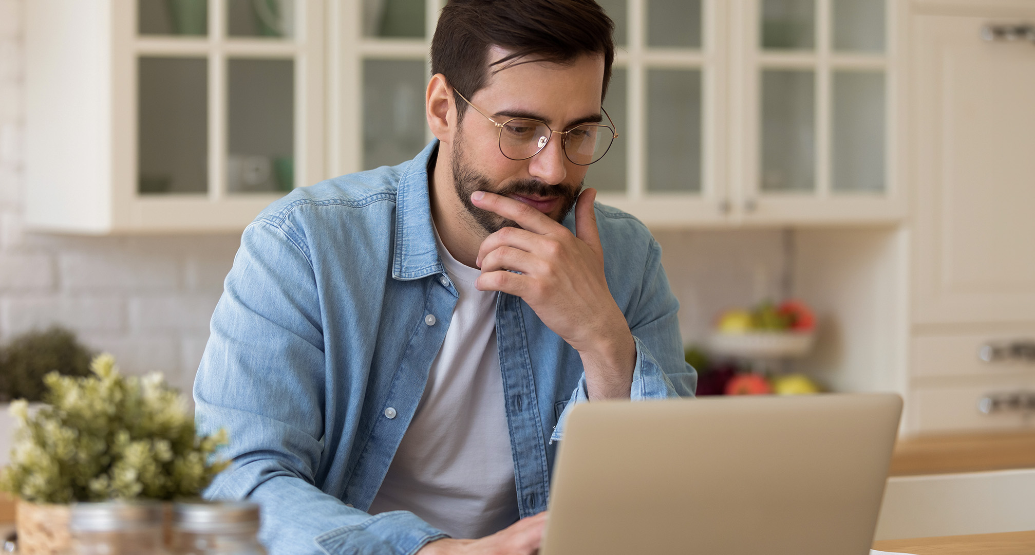 persoon met bril zit in keuken en kijkt naar laptop